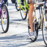 Cyclists pedalling two wheeled bikes on a road. Legs and wheels visible only, casting shadows.