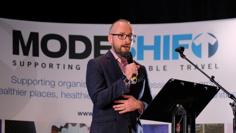 Nick Butler at lectern speaking. Modeshift display stand with logo in background.