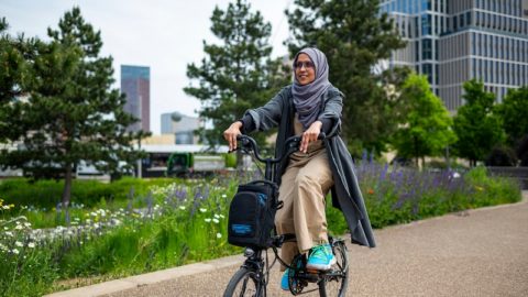 Woman rides e-bike down a path with wild flowers and city scape in the background