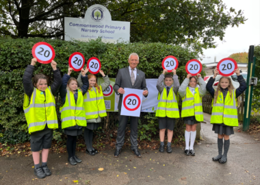 Pupils from Commonswood School wearing high viz vests and school uniform hold up 20mph signs outside the school - school sign is in the background