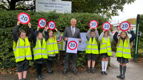 Pupils from Commonswood School wearing high viz vests and school uniform hold up 20mph signs outside the school - school sign is in the background