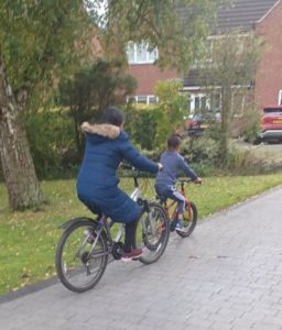 Woman and child riding bikes away from the camera down a driveway towards redbrick house. Trees and grass line the path.
