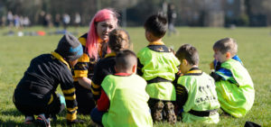 Sport England Image. Female coach sits with group of children bedside football pitch. Children wear black and yellow kits with high viz vests.