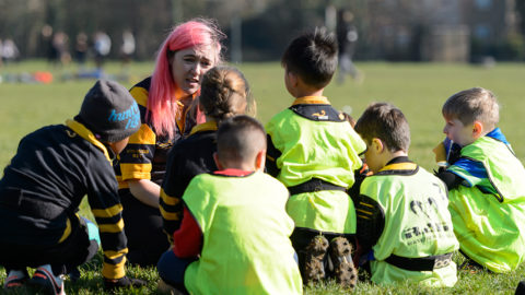 Sport England Image. Female coach sits with group of children bedside football pitch. Children wear black and yellow kits with high viz vests.