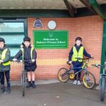 Four pupils stand with bikes, wearing high viz vests. Stand either side of the Knowsley Primary School sign outside the school building.
