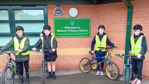 Four pupils stand with bikes, wearing high viz vests. Stand either side of the Knowsley Primary School sign outside the school building.