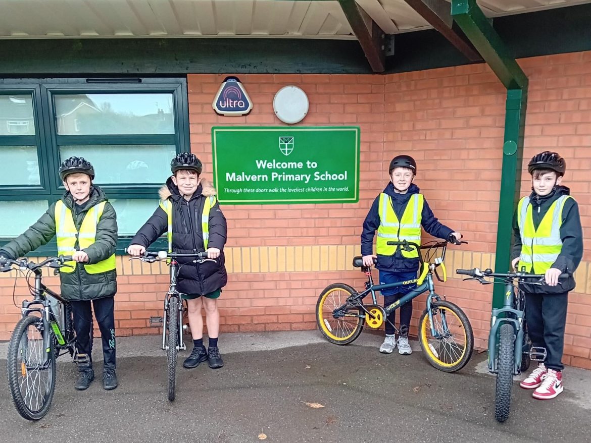 Four pupils stand with bikes, wearing high viz vests. Stand either side of the Knowsley Primary School sign outside the school building.