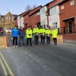 Seven people wearing high viz jackets, hats and gloves stand in a street lined by houses with school building in the background. Senior Leadership Team at Astrea Academy stand in a line by the closing of the School Street.