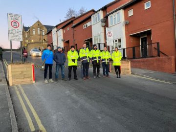 Seven people wearing high viz jackets, hats and gloves stand in a street lined by houses with school building in the background. Senior Leadership Team at Astrea Academy stand in a line by the closing of the School Street.