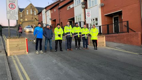 Seven people wearing high viz jackets, hats and gloves stand in a street lined by houses with school building in the background. Senior Leadership Team at Astrea Academy stand in a line by the closing of the School Street.