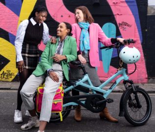 The Loud Mobility team. Three people gather around an electric bike. Wearing bright pink, green and blue clothes. The background has a wall of colourful graffiti.