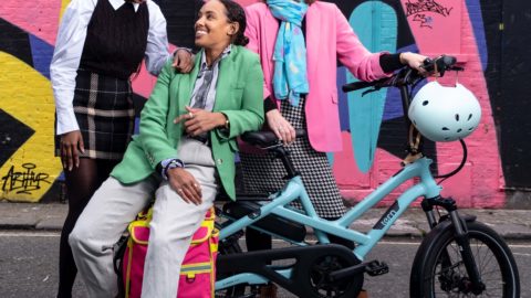The Loud Mobility team. Three people gather around an electric bike. Wearing bright pink, green and blue clothes. The background has a wall of colourful graffiti.