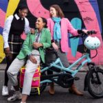 The Loud Mobility team. Three people gather around an electric bike. Wearing bright pink, green and blue clothes. The background has a wall of colourful graffiti.
