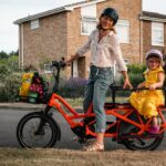 Caz Conneller poses with e-bike with little girl wearing a yellow dress seated in child seat at the back of the bike. Residential street in the back ground with house and bushes surrounding.