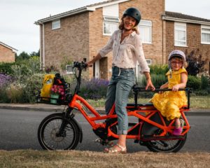 Caz Conneller poses with e-bike with little girl wearing a yellow dress seated in child seat at the back of the bike. Residential street in the back ground with house and bushes surrounding.