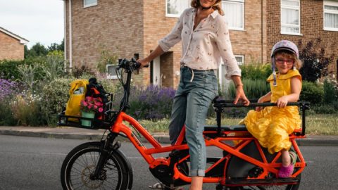 Caz Conneller poses with e-bike with little girl wearing a yellow dress seated in child seat at the back of the bike. Residential street in the back ground with house and bushes surrounding.