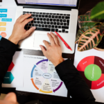 persons hands at laptop surrounded by papers with pie charts and diagrams on.