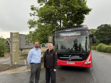 Two people stand by entrance to Harlow Carr, red bus with 'Harrogate' visible in background.