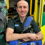Andy sits by ambulance wearing his green paramedic uniform, smiling at the camera