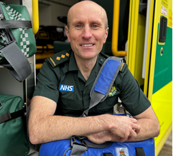 Andy sits by ambulance wearing his green paramedic uniform, smiling at the camera