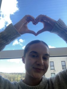 Steph makes a heart shape with her hands above her head. Blue sky with clouds in background.