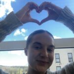 Steph makes a heart shape with her hands above her head. Blue sky with clouds in background.