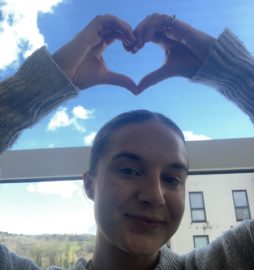 Steph makes a heart shape with her hands above her head. Blue sky with clouds in background.
