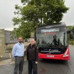 Two men stand in front of a red single deck bus posing for the camera. Harlow Carr sign to their left at the entrance.