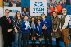 Four children stand in the centre with two adults on either side of the group. The shildren wear blue and grey school uniform. A boy on the far right holds a star shaped award.