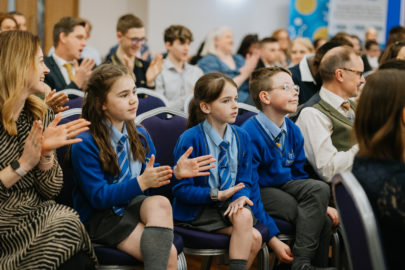 Group people seated in rows. Closest to the camera are three school children. They are wearing blue school jumpers with grey trousers and skirts/long socks.