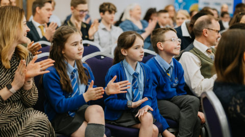 Group people seated in rows. Closest to the camera are three school children. They are wearing blue school jumpers with grey trousers and skirts/long socks.