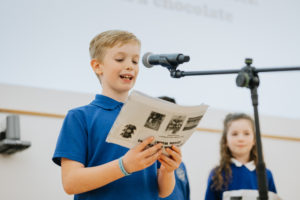 Two school children stand in front of microphones. Boy wearing blue t-shirt reads from a booklet.