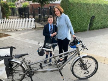 Mum and daughter stand next to tandem bike outside of school building.