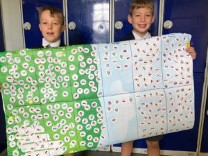 Two school students in school uniform stand holding large sticker chart, covered with multicoloured stickers.