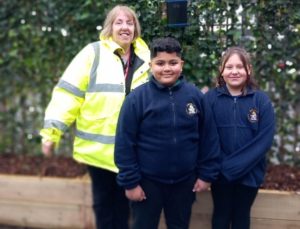 Two school pupils in blue school uniform stand with woman in high viz yellow coat. A fence covered with greenery is behind them.