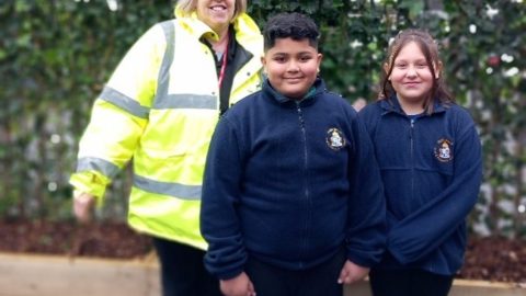 Two school pupils in blue school uniform stand with woman in high viz yellow coat. A fence covered with greenery is behind them.