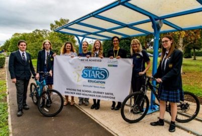 School students in uniform hold a large Modeshift STARS Banner. The group of eight stand beside bicycle shelter, two pose with bikes