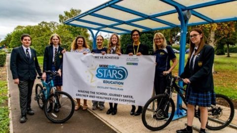 School students in uniform hold a large Modeshift STARS Banner. The group of eight stand beside bicycle shelter, two pose with bikes