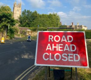 Red sign reads: 'Road ahead closed'. Empty road in background. Church and green trees in the distance.