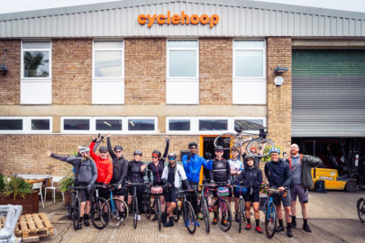 Group of cyclists stand with bikes outside a brick building with large windows. Sign reads: Cyclehoop