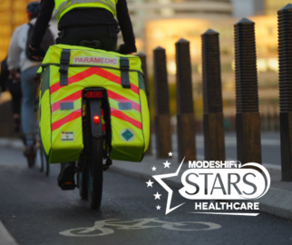 Paramedic riding bike away from the camera. Yellow equipment bag with text: Paramedic written in bright red. A white bicycle is painted in white on the road beneath. STARS Healthcare logo in bottom right corner.