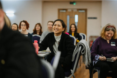 Group of people seated in a meeting, camera focus is on woman wearing black jacket smiling.