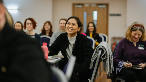 Group of people seated in a meeting, camera focus is on woman wearing black jacket smiling.