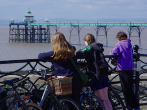 Three girls look over railing out to sea. A pier is in the distance. They have bikes stacked to their left. Image from North Somerset Council.