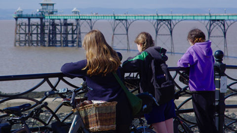 Three girls look over railing out to sea. A pier is in the distance. They have bikes stacked to their left. Image from North Somerset Council.