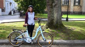 Woman stands with ebike. Wearing cycle helmet and smiling at the camera. Tress and green grass in the background. Image from Sustrans.