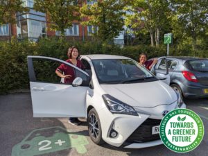 Two women in scrubs uniform stand by white car smiling.