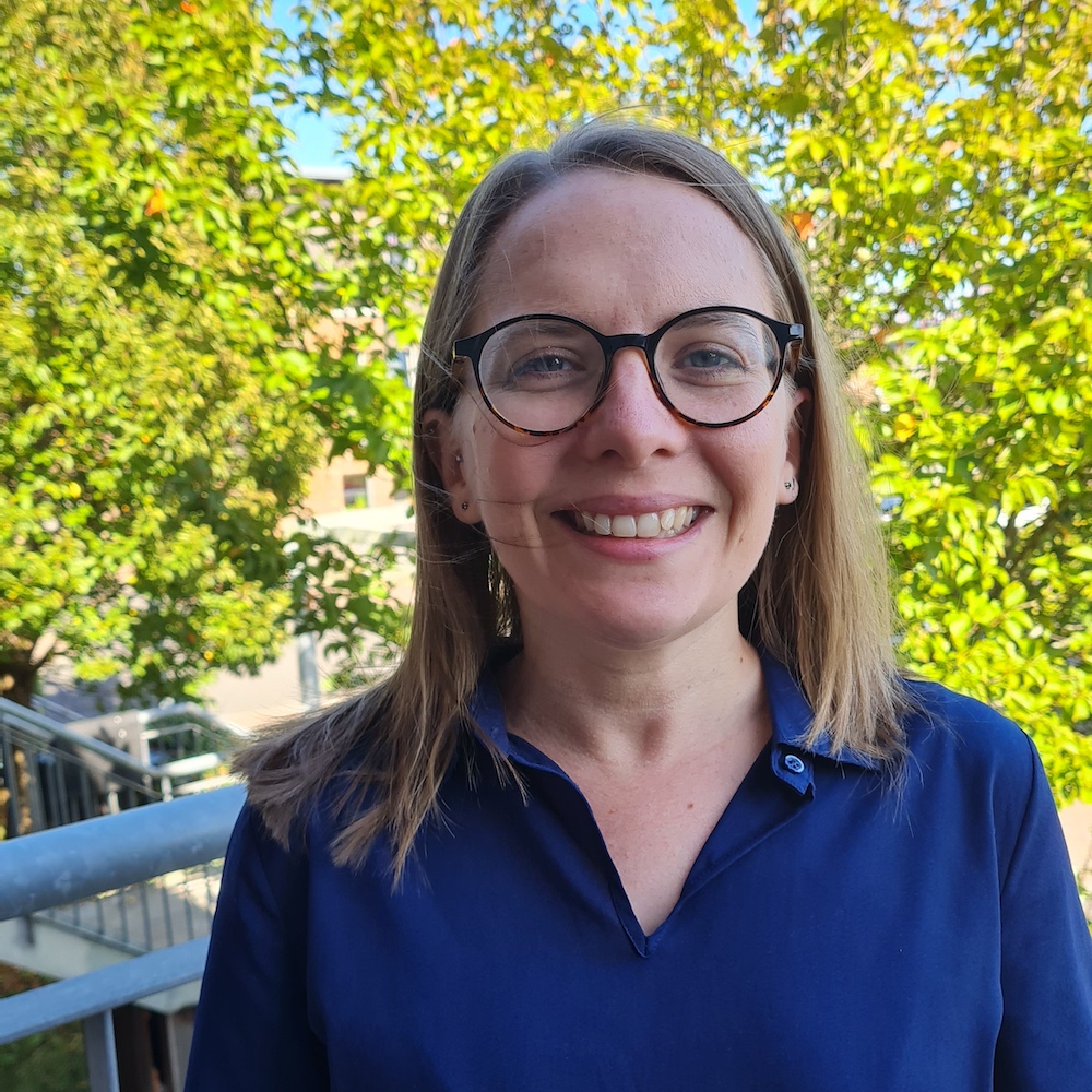 Corrine smiles at the camera. She wears a blue top and black framed glasses. Green trees in the background.