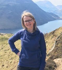 Woman stands with hand on hip, wearing blue top and glasses. In the background there is a mountainous landscape with a lake in view.