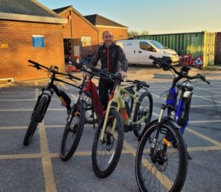 Man stands with four e-bikes in a row. Bick building a white van in background.